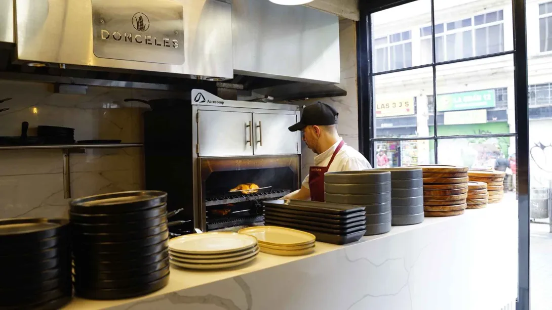 Un cocinero preparando alimentos frente al horno en una cocina industrial moderna rodeado de vajilla apilada.