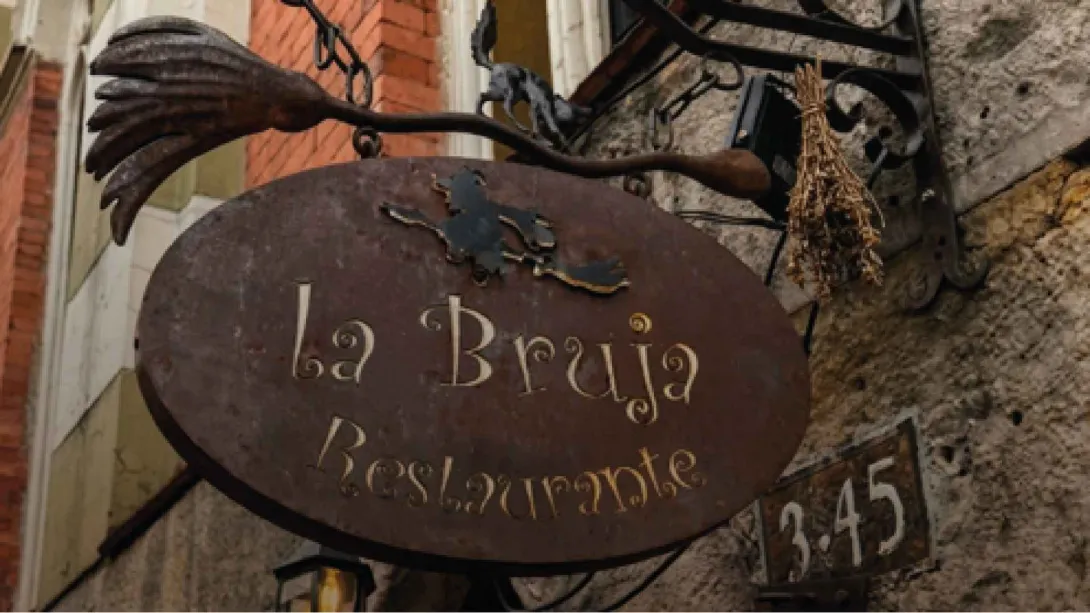 Un letrero ovalado de metal oxidado colgando, con el texto dorado "la Bruja Restaurante" y una figura de bruja.