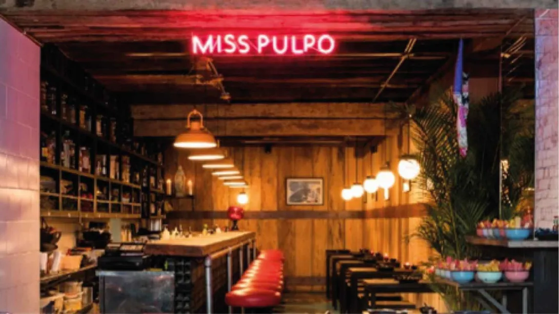 Barra de bar en madera con bancos rojos bajo un letrero neón de Miss Pulpo y cálidas lámparas colgantes.