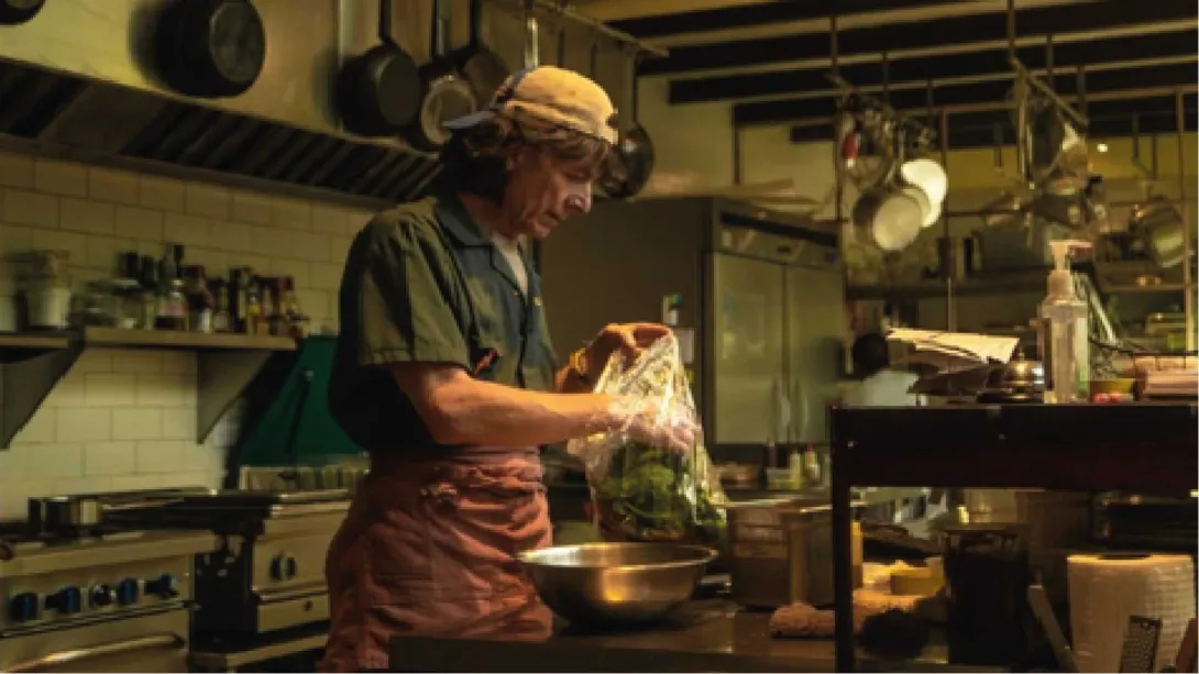 Un chef con una gorra de béisbol y un delantal de cocina prepara verduras en una bolsa de plástico en una cocina rústica.