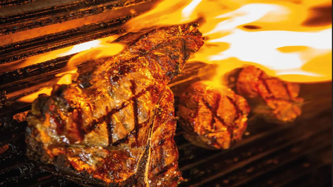 Grueso corte de carne T-bone asándose a las brasas sobre una parrilla metálica con llamas de fuego alrededor.