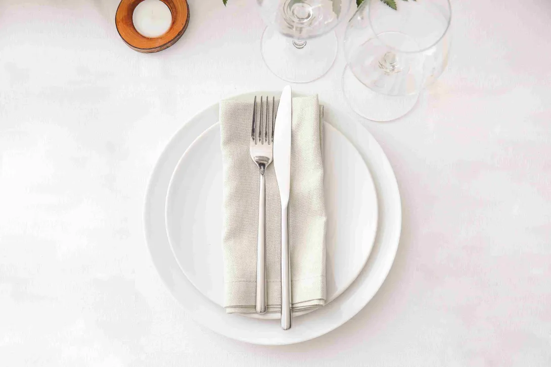 Mesa de restaurante con plato blanco, cubiertos y servilleta, preparada para recibir a los comensales.