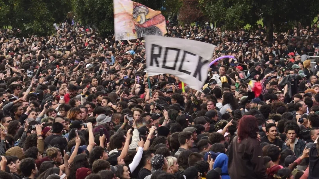 Multitud de personas en un concierto al aire libre, levantando manos y banderas, con un cartel que dice “ROCK” en medio del público.