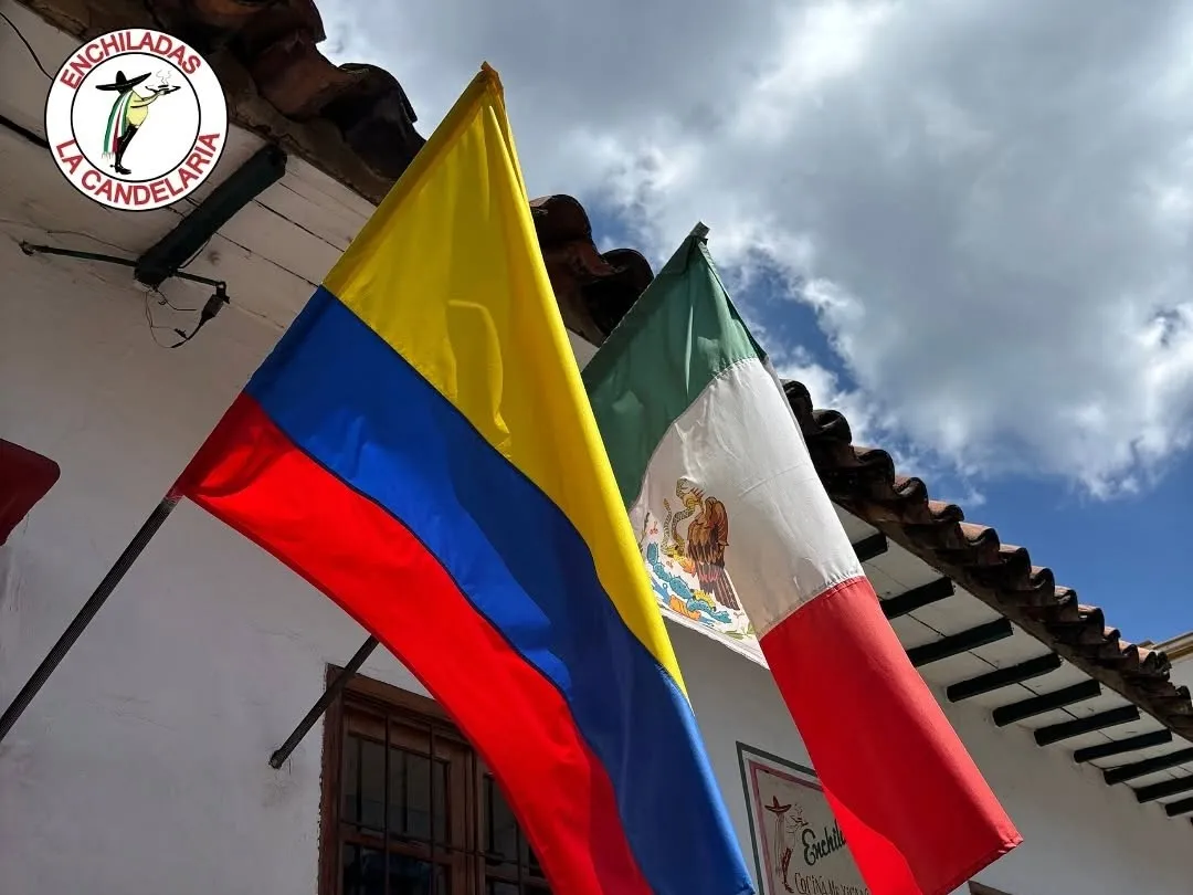 Banderas de Colombia y México ondean frente a la fachada colonial de Enchiladas La Candelaria, bajo un cielo parcialmente nublado.