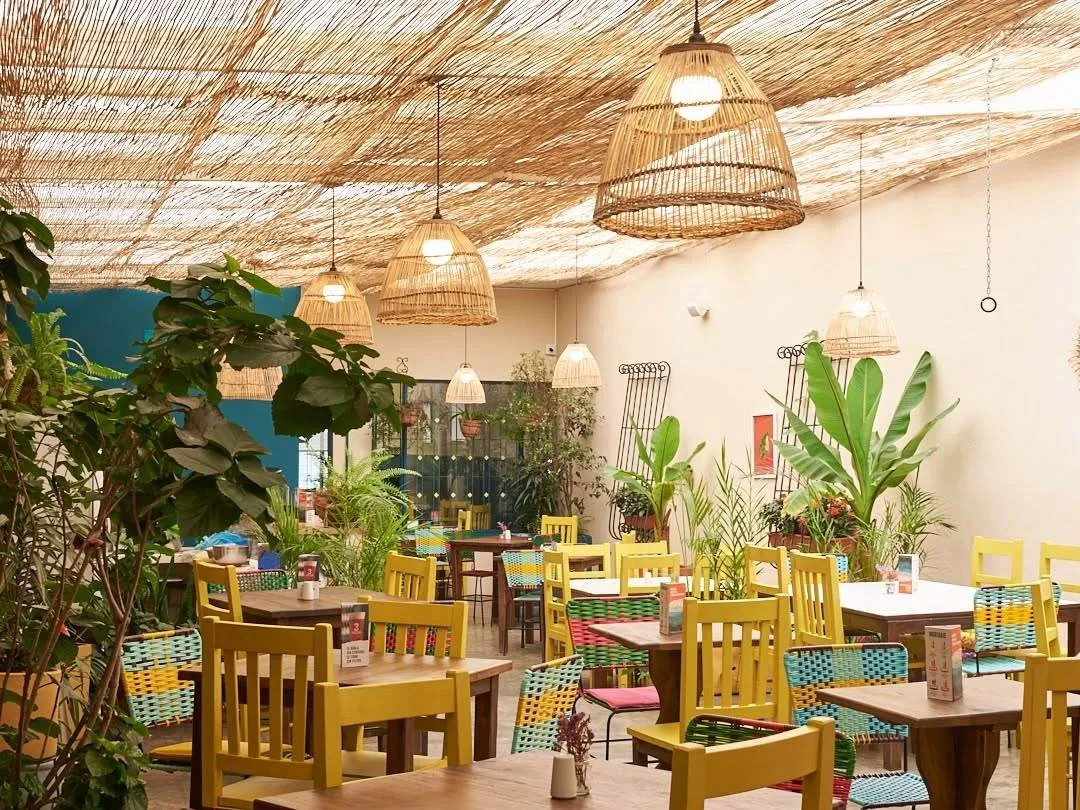 Interior del restaurante con mesas de madera, sillas coloridas, abundante vegetación y lámparas de mimbre que crean un ambiente cálido y acogedor.