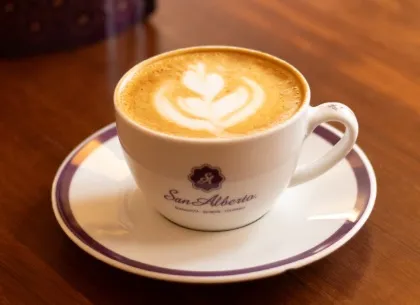 Taza de café San Alberto con delicada espuma en forma de corazón, servida sobre un elegante plato en una mesa de madera.