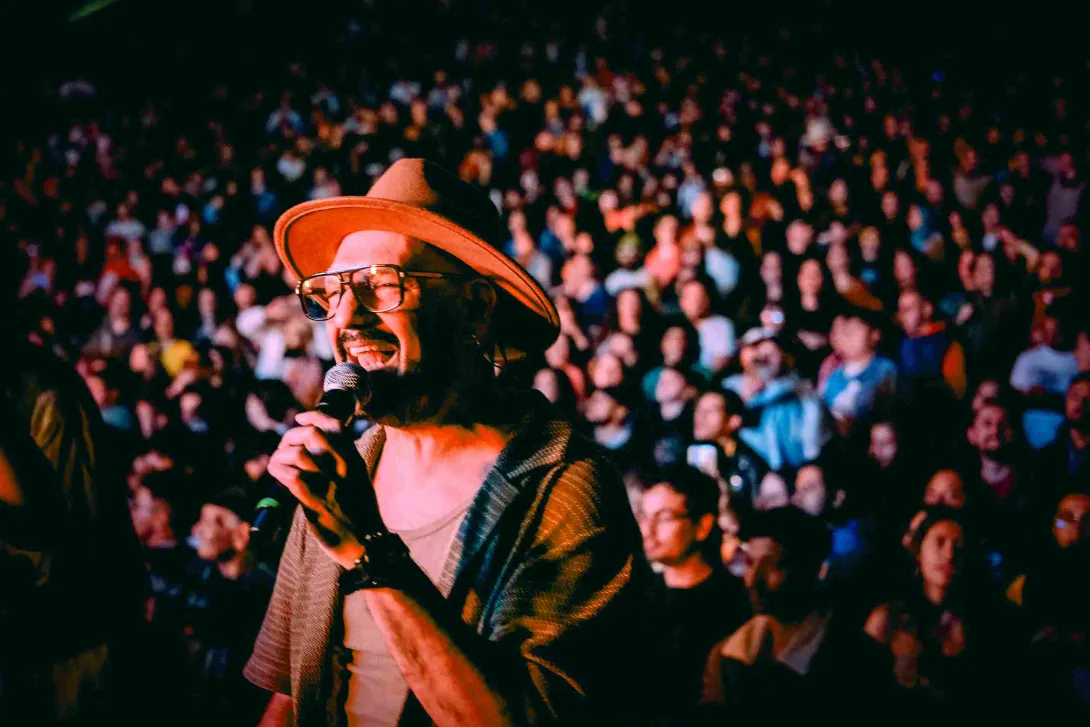 Cantante con sombrero y gafas interpreta con micrófono frente a una multitud en un concierto nocturno.
