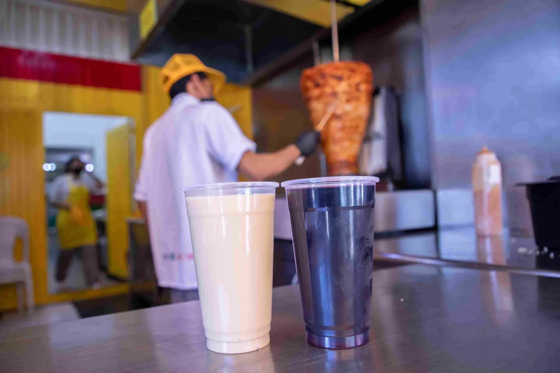  Dos bebidas servidas en vasos y un trompo de carne al pastor al fondo del restaurante.