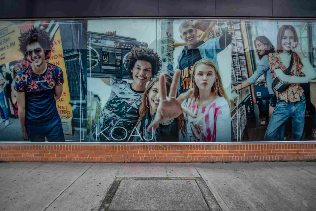 Un gran mural publicitario ocupa el escaparate de la tienda KOAJ, con imágenes de jóvenes y una estética urbana y colorida que refleja el espíritu moderno de la marca. La fachada combina los ventanales con una base de ladrillo característica de la edificación.