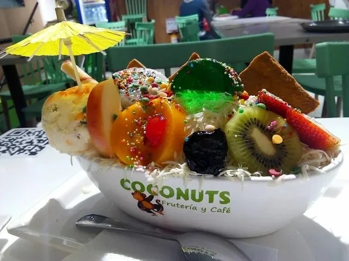 Copa de frutas tropicales con helado, gelatina, coco rallado, grageas y salsa dulce, servida en un recipiente de la frutería.