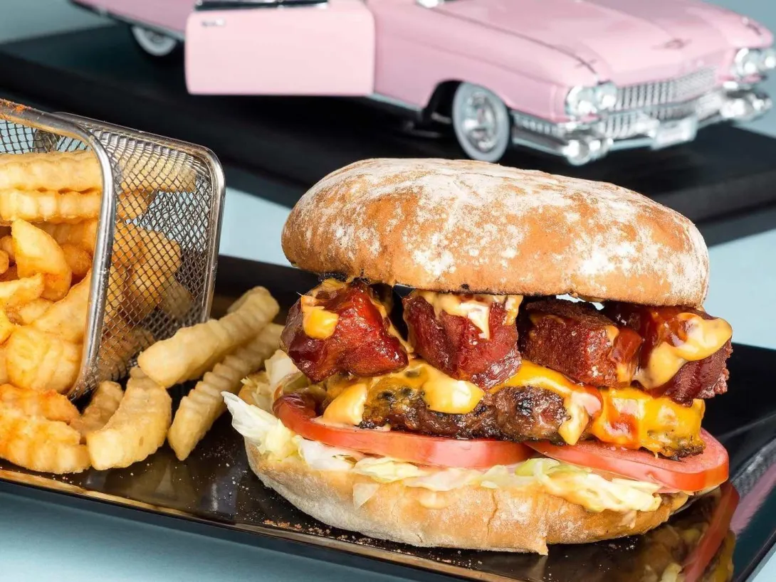  Hamburguesa doble con queso, tocino y salsas, acompañada de papas, en una presentación inspirada en los clásicos años 50.