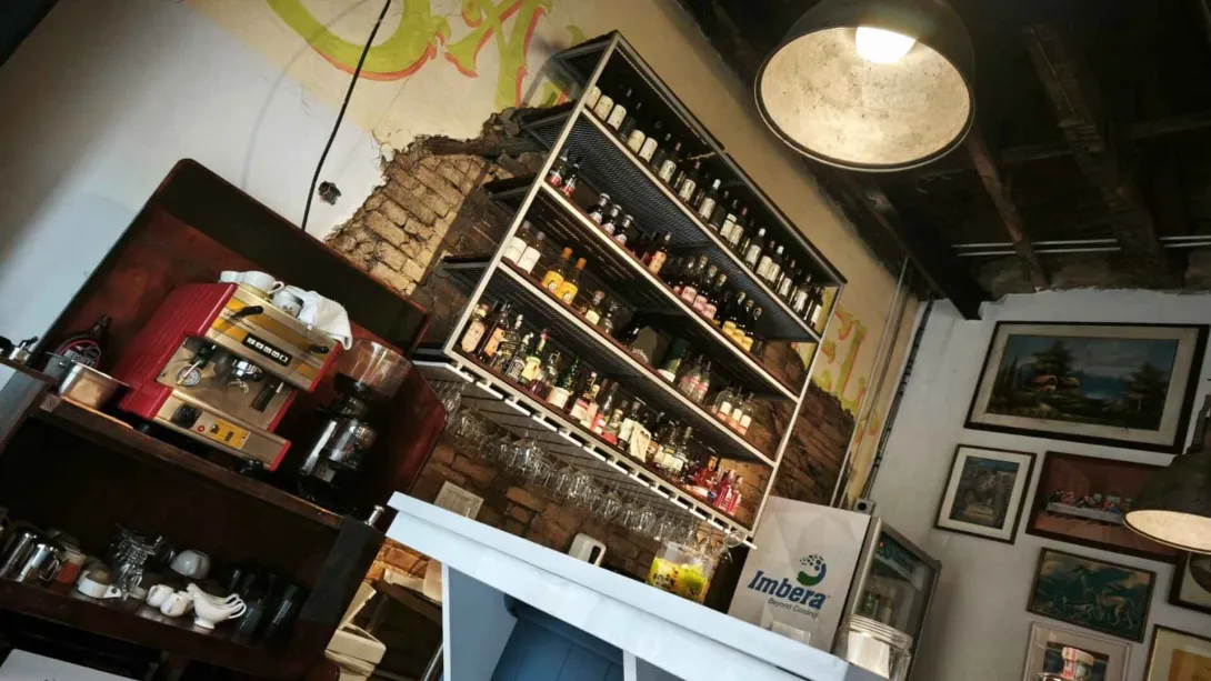 Barra de bar con repisas de licores iluminadas, máquina de café espresso roja y pared de ladrillo expuesto. 