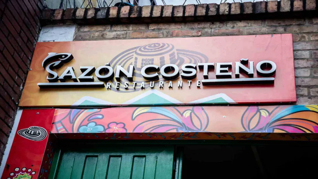 Letrero del restaurante Sazón Costeño sobre fachada de ladrillo con ilustración de sombrero vueltiao y colores.
