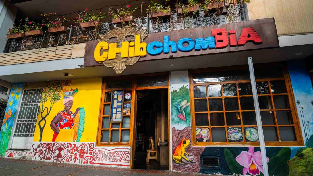 Fachada colorida con el letrero Chibchombia, murales artísticos folclóricos y ventanales con marcos de madera.