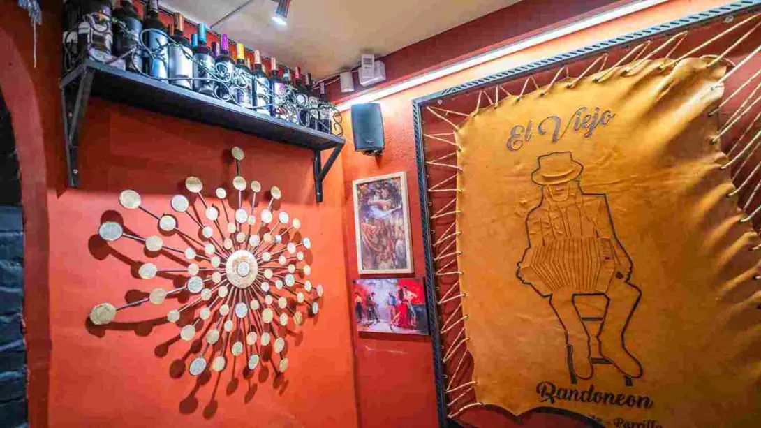 Pared roja decorada con adornos metálicos, botellas de vino en estante y tapiz de cuero con el logo de El Viejo.