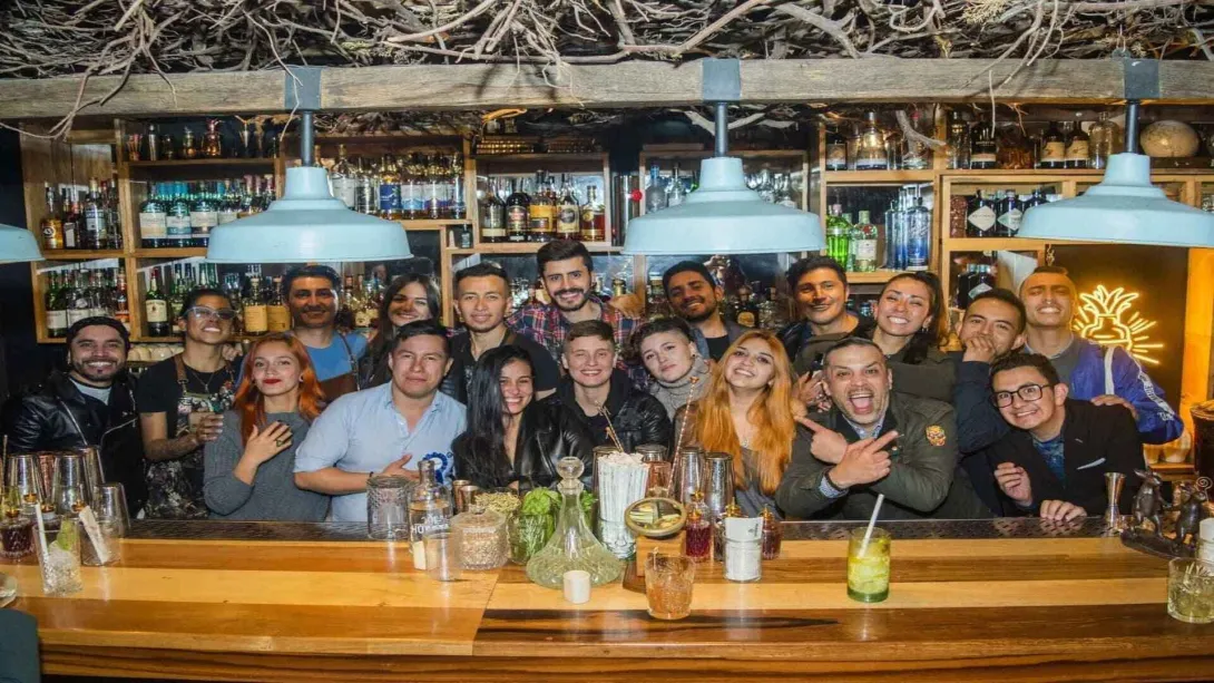 Un grupo diverso de jóvenes sonrientes posa detrás de una barra de bar bien surtida, con bebidas y herramientas de coctelería.