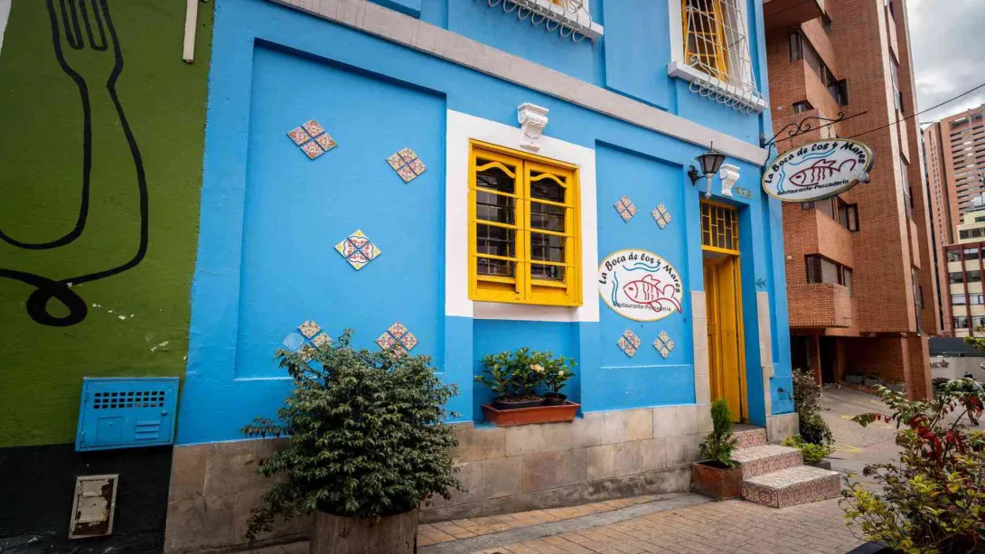 Edificio con fachada azul, ventana y puerta amarillas. El letrero circular del restaurante y plantas decoran la entrada.