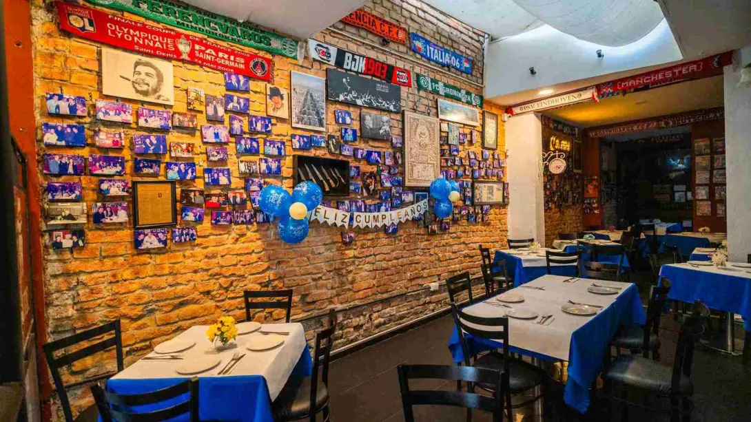 Comedor con mesas vestidas de mantel azul y blanco, frente a una pared de ladrillo repleta de fotografías, bufandas de fútbol y decoración de cumpleaños.