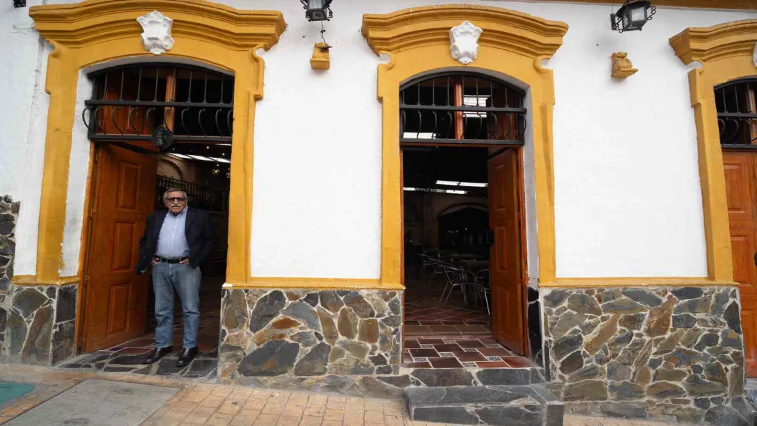 Hombre mayor con gafas y saco oscuro parado en la puerta de entrada de una casona colonial con detalles amarillos.