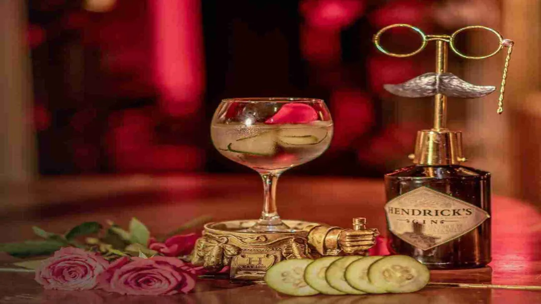 Un elegante cóctel de ginebra con pepino y pétalos de rosa, acompañado de una botella de Hendrick's Gin y adornos vintage, sobre un fondo rojo romántico.