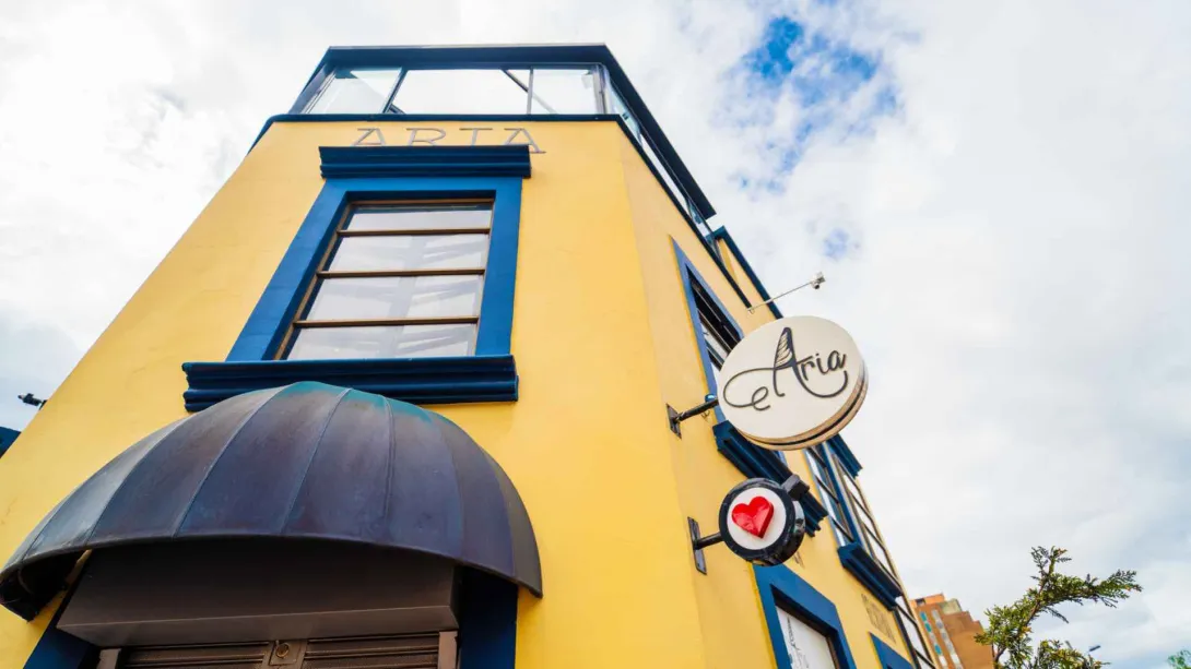 Fachada de un edificio amarillo de esquina con marcos de ventanas azules, marquesina oscura y letreros circulares de la marca Aria.