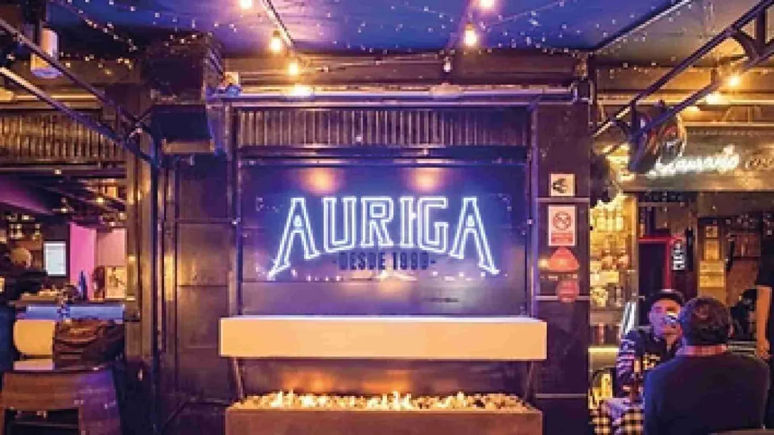 Letrero luminoso de "AURIGA DESDE 1998" sobre una chimenea encendida en el interior del bar. 