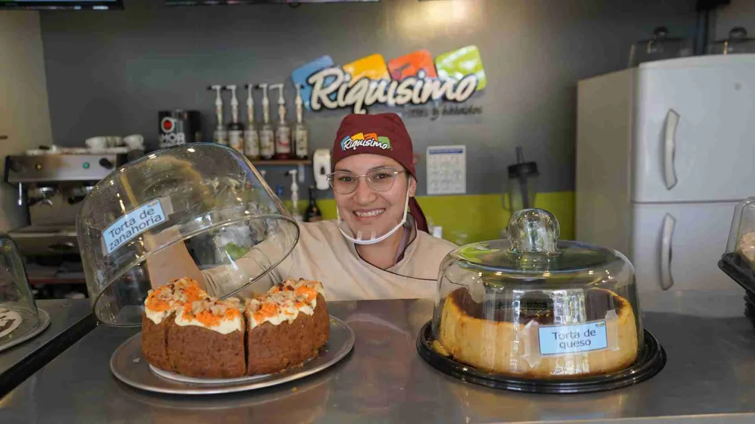 Pastelera sonriente con uniforme detrás del mostrador sosteniendo una campana de vidrio sobre una torta de zanahoria. 