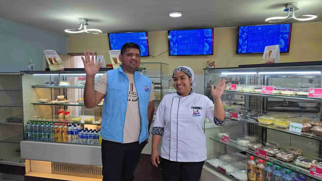  Un hombre con chaleco azul y una mujer con uniforme de chef sonríen y saludan frente a vitrinas llenas de postres en un establecimiento llamado "La Torre de los Postres".