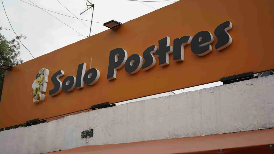 Letrero de fachada de color naranja con letras negras y blancas en relieve que dice Solo Postres junto a un panal. 