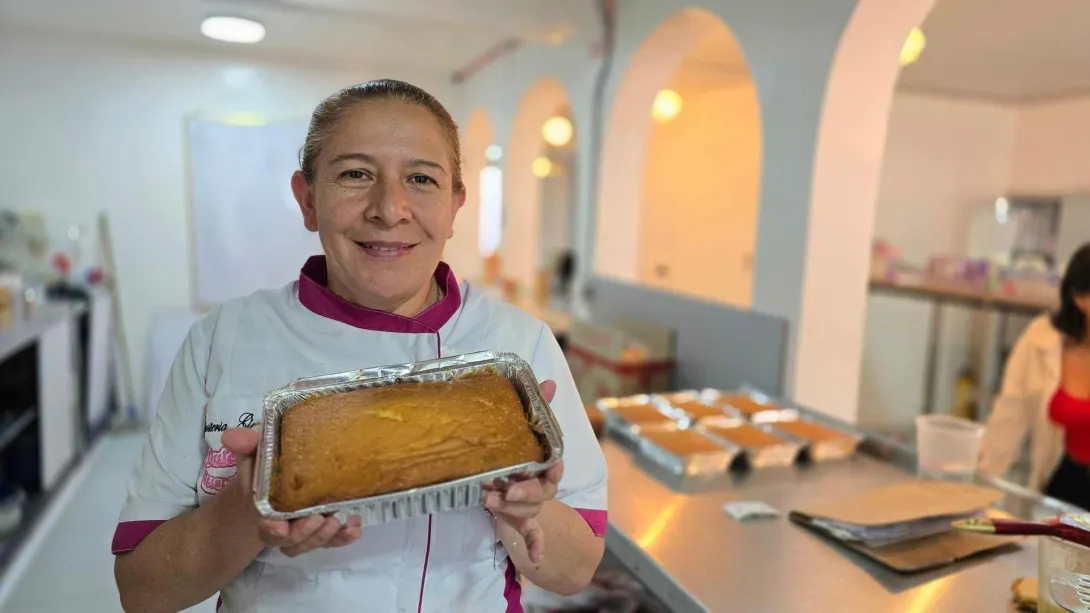 Mujer repostera sonríe mientras sostiene una bandeja de aluminio con un ponqué recién horneado. 