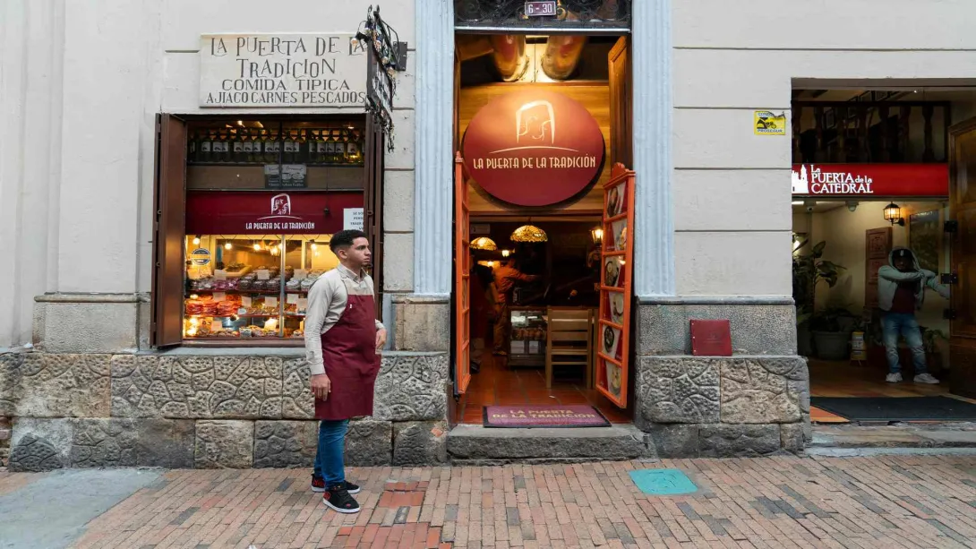 Fachada del restaurante La Puerta de la Tradición con un mesero de delantal rojo parado en la entrada. 