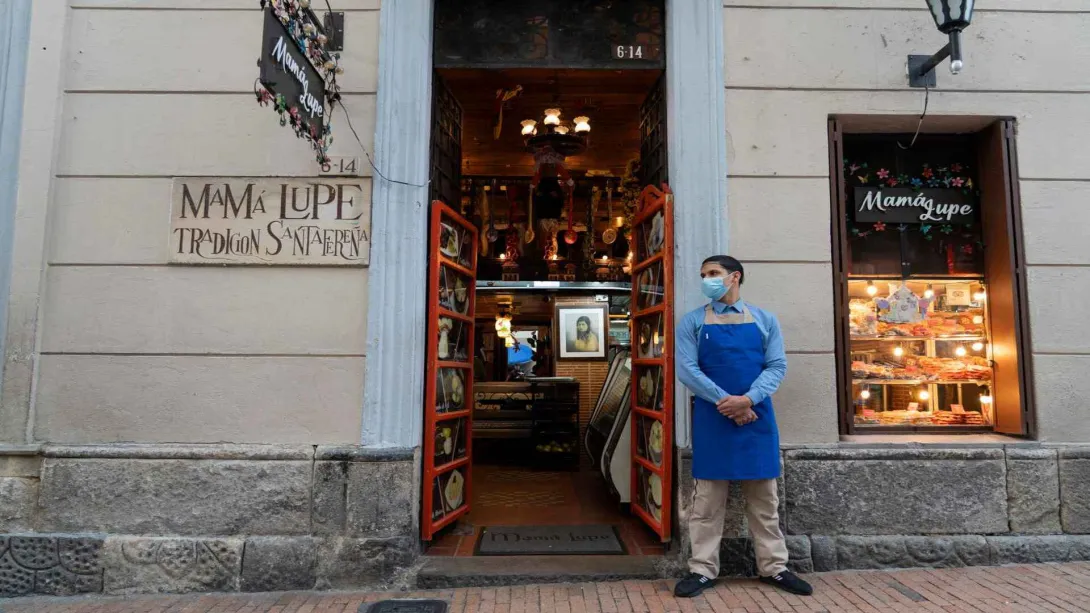  Empleado con tapabocas y delantal azul en la puerta de entrada de una edificación colonial con letreros tradicionales. 