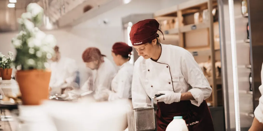 Chef de Vapiano preparando alimentos en la cocina, con otros integrantes del equipo trabajando al fondo.
