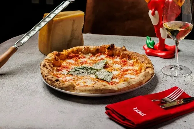 Pizza napolitana de masa artesanal, cubierta con tomate, mozzarella y albahaca, acompañada de queso parmesano y una copa de vino.