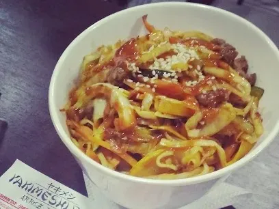 Plato de fideos salteados con carne, vegetales y salsa, acompañado de ajonjolí, servido en un tazón blanco.