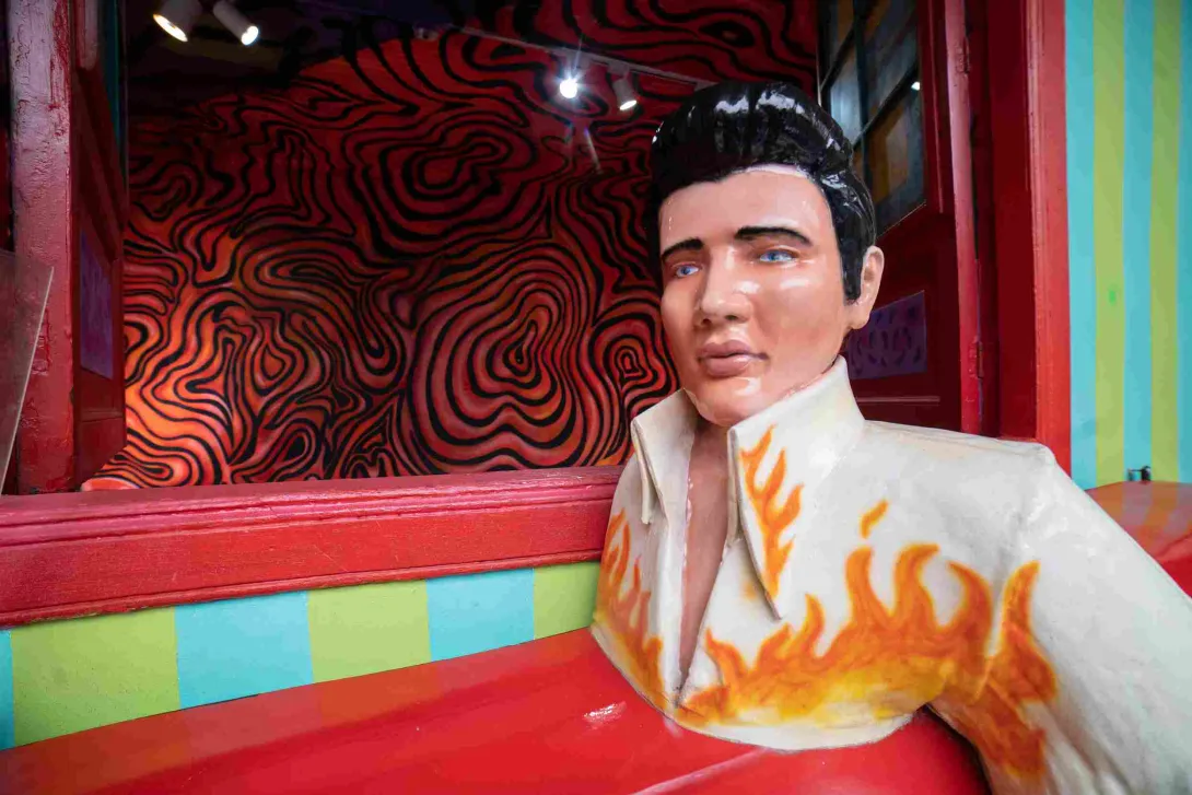 Escultura de una figura masculina con apariencia de estrella de rock, vestida con una chaqueta blanca decorada con llamas naranjas, ubicada en uno de los coloridos escenarios del Museo de la Selfie en Bogotá. Detrás se observa una pared con diseños psicodélicos en tonos rojos y negros.