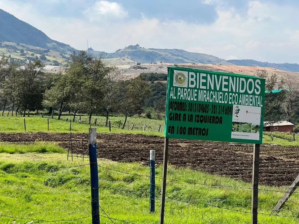 Un aviso verde da la bienvenida al Parque Mirachuelo Ecoambiental e indica el desvío para llegar, rodeado de praderas, cultivos y montañas que caracterizan el paisaje rural de Ciudad Bolívar.