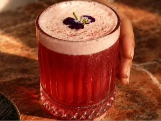 Cóctel de color rojo intenso, servido en vaso bajo y decorado con espuma y una flor comestible.