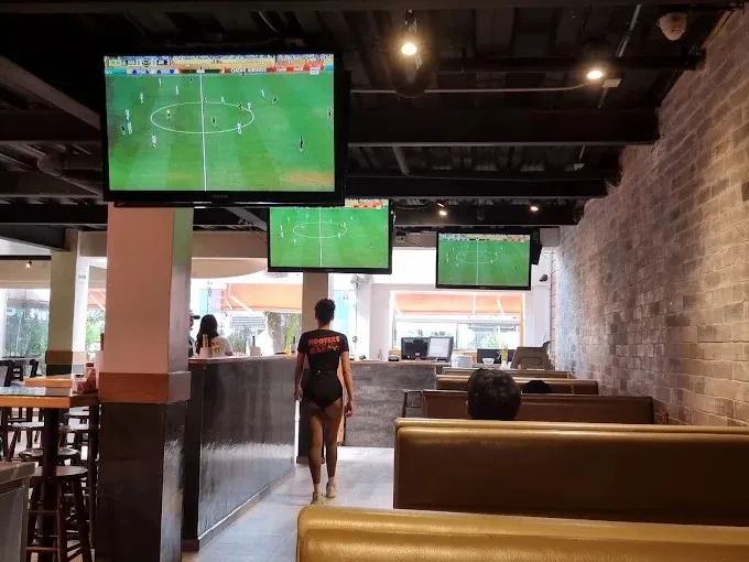 Interior de Hooters Zona T con pantallas que transmiten un partido de fútbol, mesas y cómodas zonas para disfrutar del ambiente deportivo.