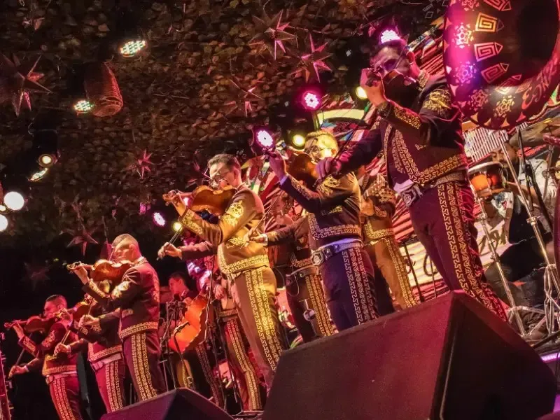 Grupo de mariachis interpretando música en vivo sobre un escenario, con iluminación colorida y ambiente festivo.